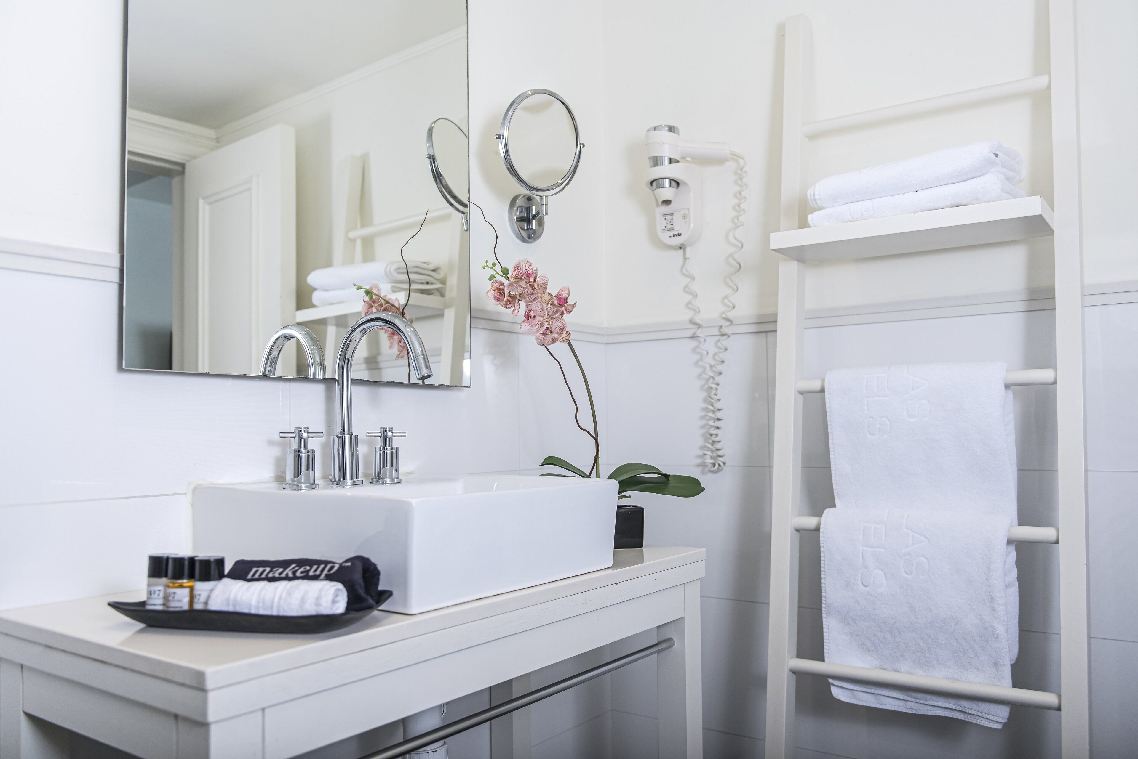 A modern bathroom with a white sink, two faucets, a large mirror, a hairdryer, a towel rack, and a small potted plant.