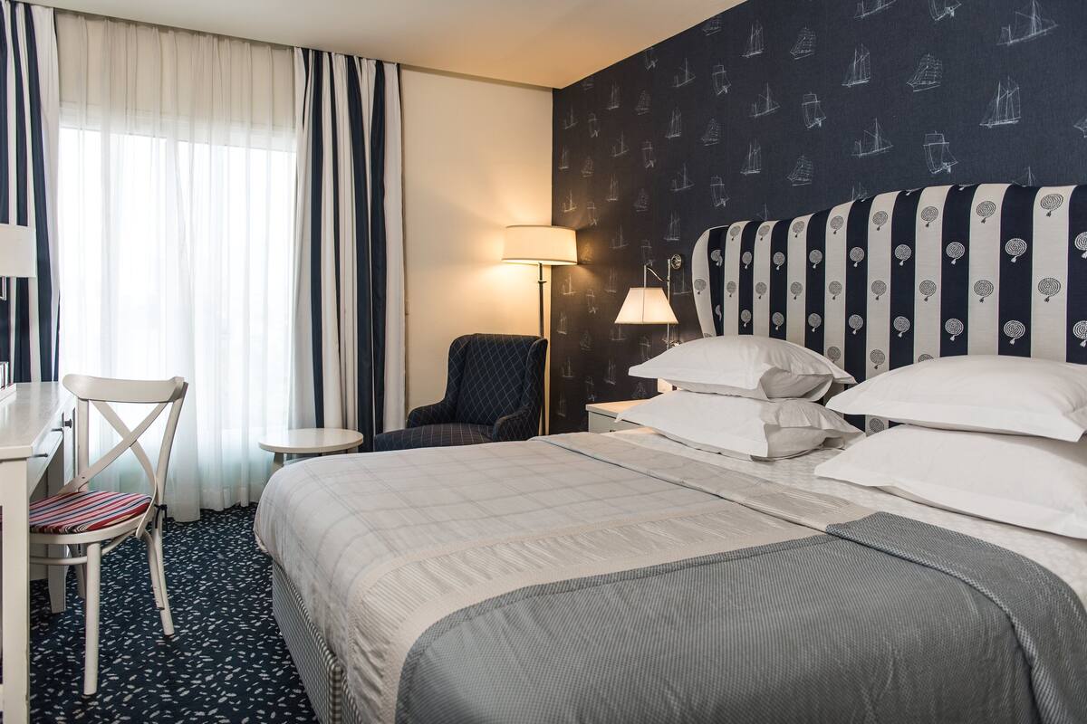 A hotel room with a large bed, a desk with a chair, a small table, and a lamp.