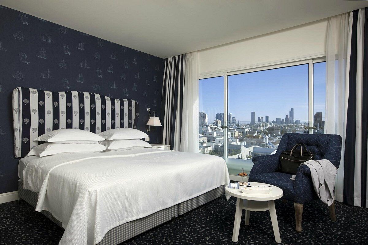 Ein Hotelzimmer mit einem großen Bett, einem Sessel, einem kleinen Tisch und Blick auf die Stadt.
