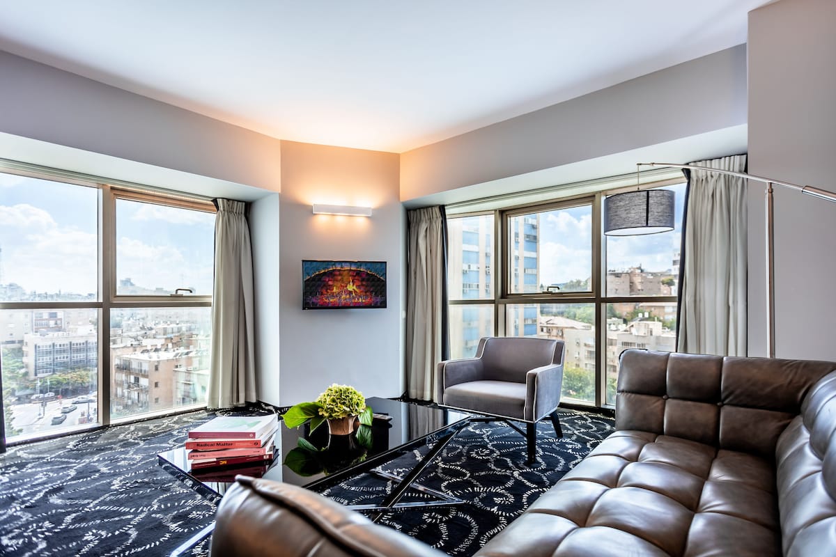 A modern living room with a leather sofa, a chair, a coffee table, and a view of the city through large windows.