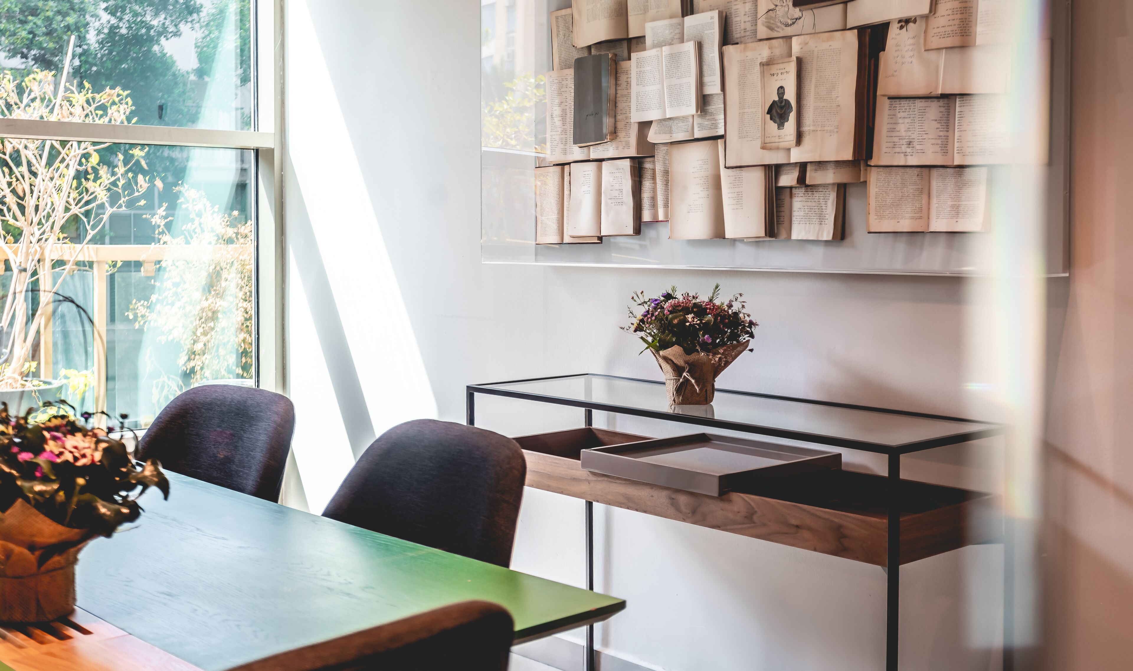 A modern room with a glass table, two chairs, a vase with flowers, and a wall decorated with papers and books.