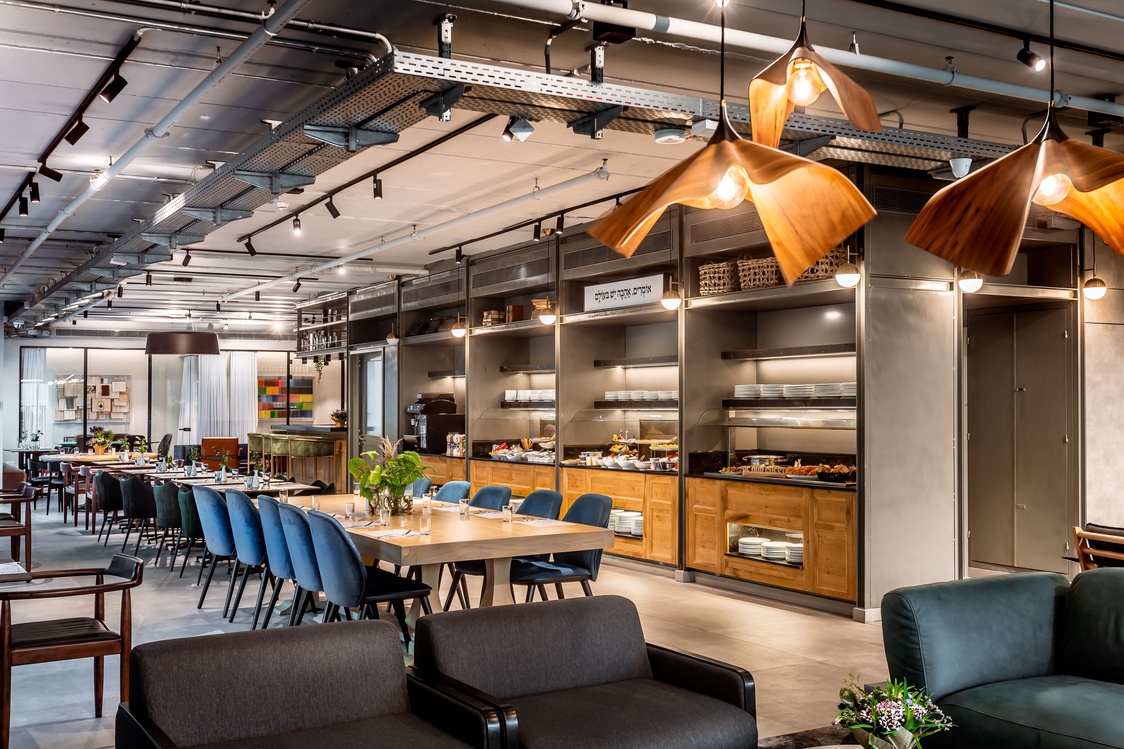 Un área de comedor moderna con una mesa larga, sillas azules y un sofá. Se observan estanterías con alimentos y una zona de cocina al fondo.