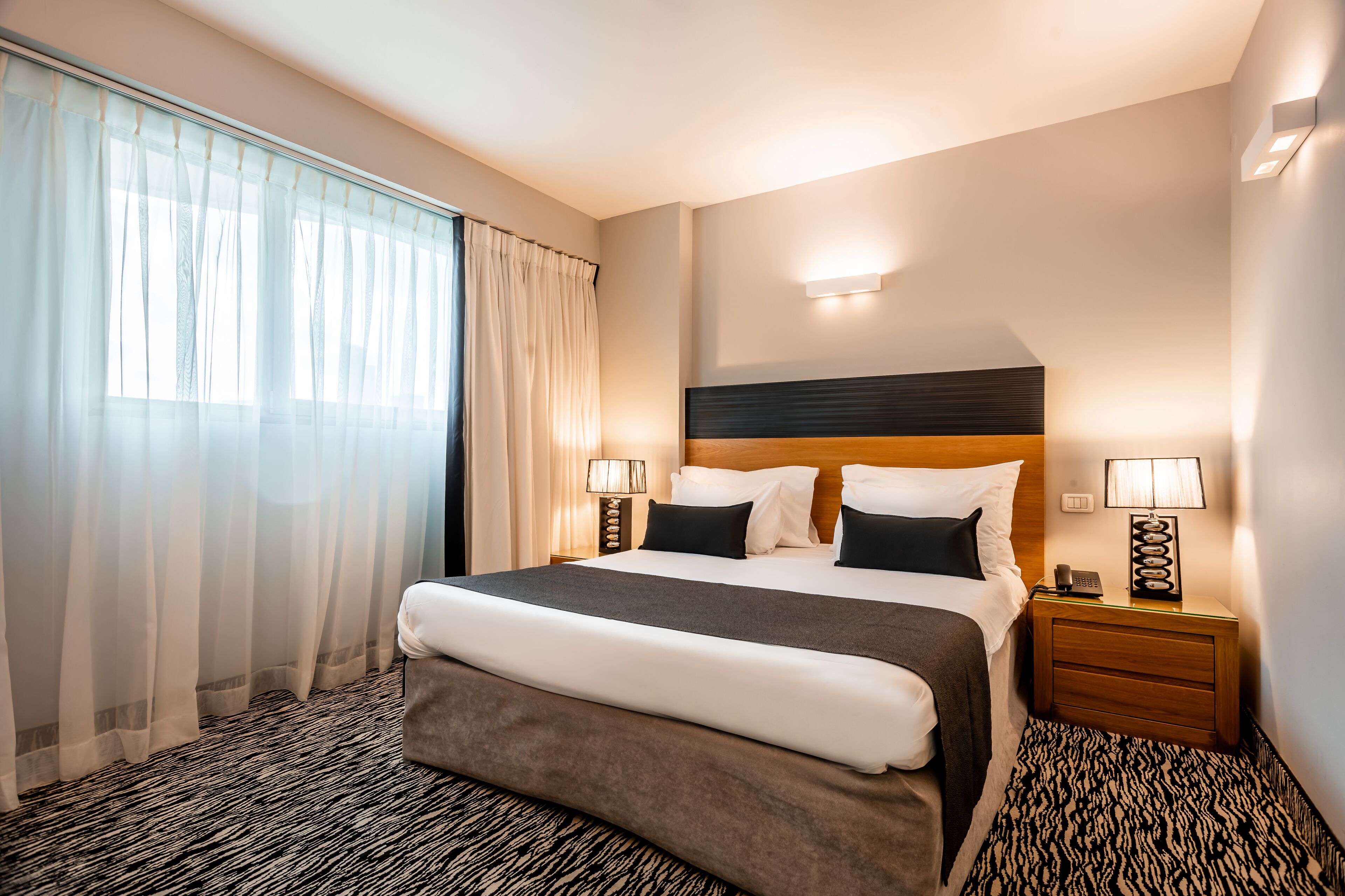 A hotel room with a large bed, two bedside tables with lamps, a headboard, and a window with sheer curtains.