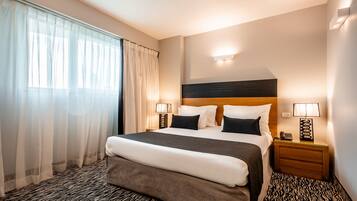 A hotel room with a large bed, two bedside tables with lamps, a headboard, and a window with sheer curtains.