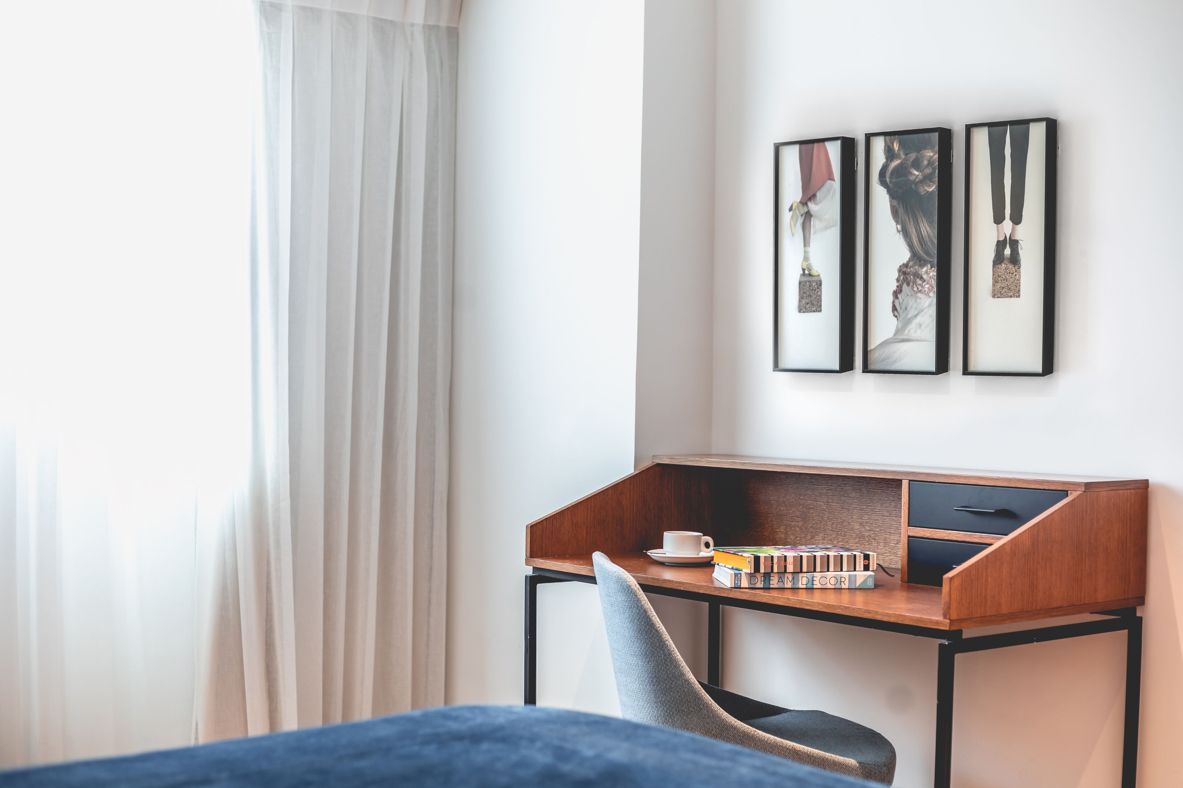 A hotel room with a wooden desk, a chair, a bed, and a window with curtains. There are framed pictures on the wall.