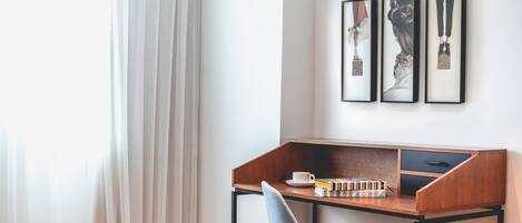 Habitación de hotel con un escritorio de madera, una silla, una cama y una ventana con cortinas. Hay cuadros enmarcados en la pared.