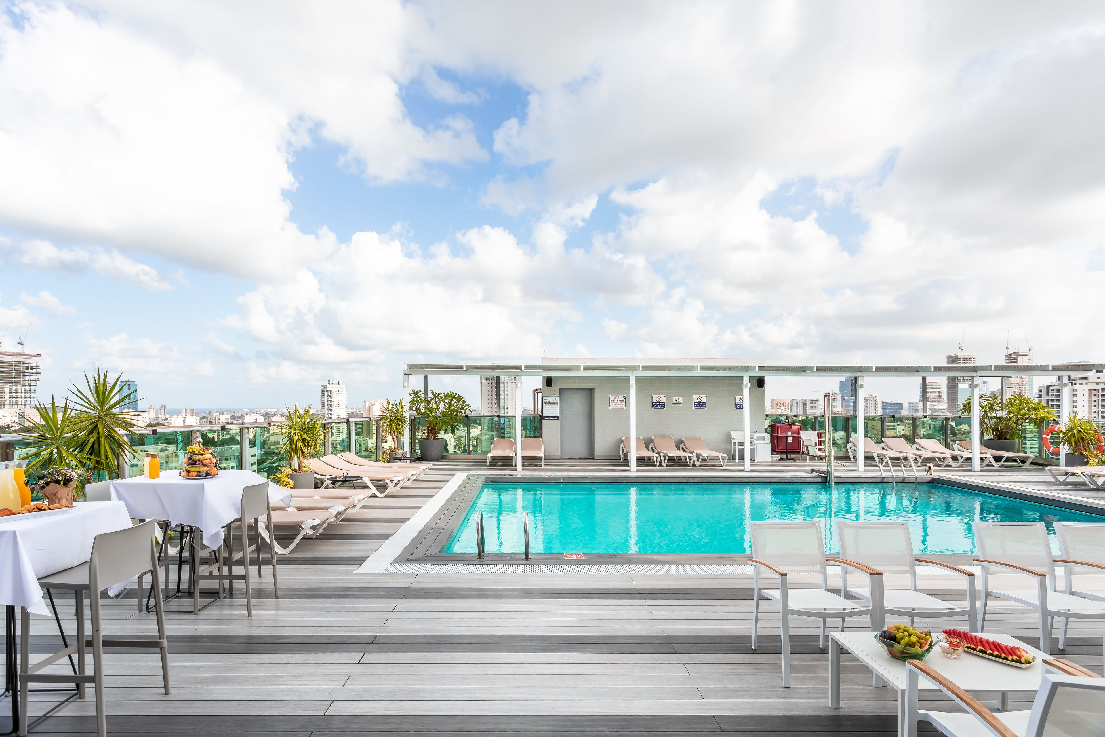 A rooftop pool area with lounge chairs, a dining table, and a view of the city skyline.