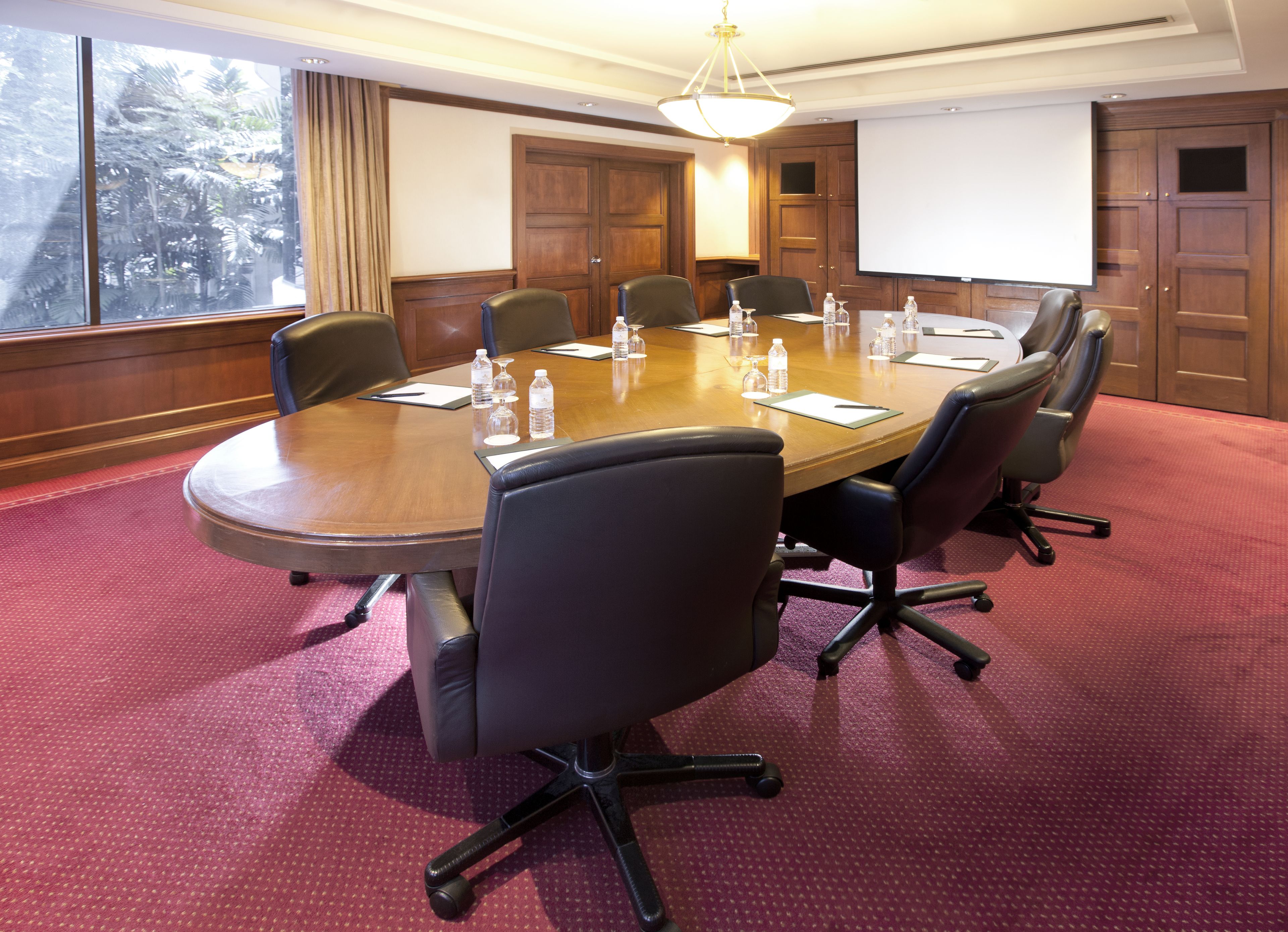 A conference room with a large oval table, six chairs, water bottles, and a projector screen.
