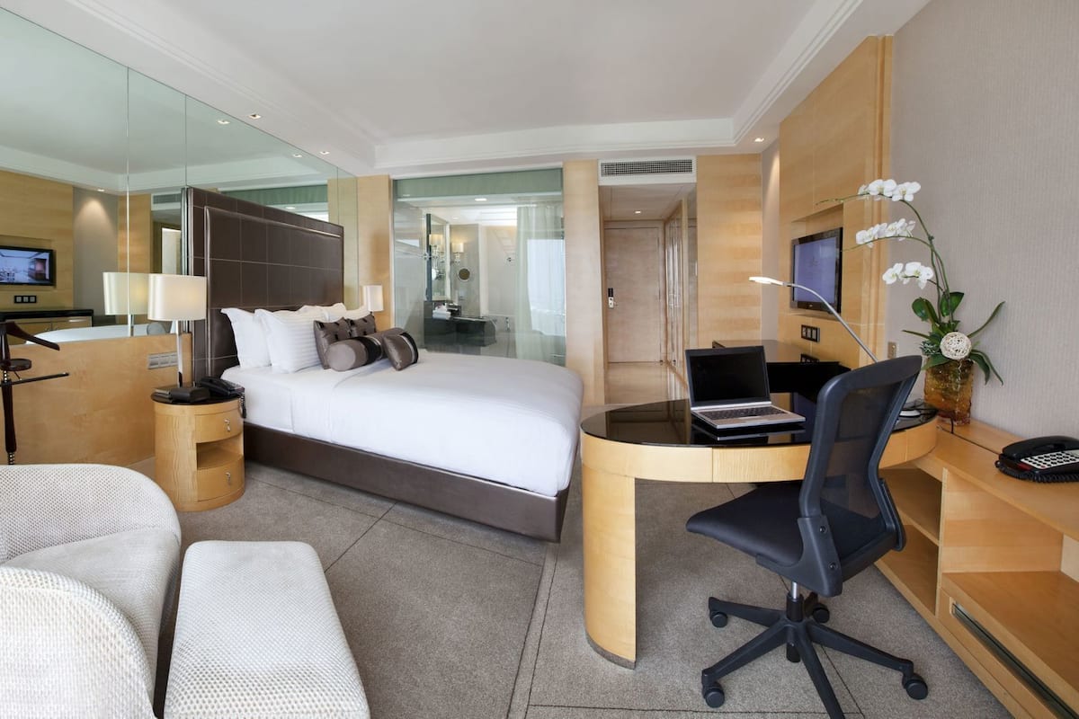 A hotel room with a bed, a desk with a laptop, a chair, a bedside table, and a mirror.