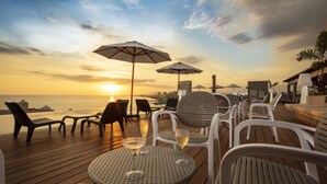 Eine Holzterrasse mit Liegestühlen und Sonnenschirmen mit Blick auf einen Sonnenuntergang über dem Meer.