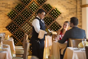 Un restaurante con mesas dispuestas para comer, una estantería con botellas de vino al fondo y un camarero sirviendo vino.