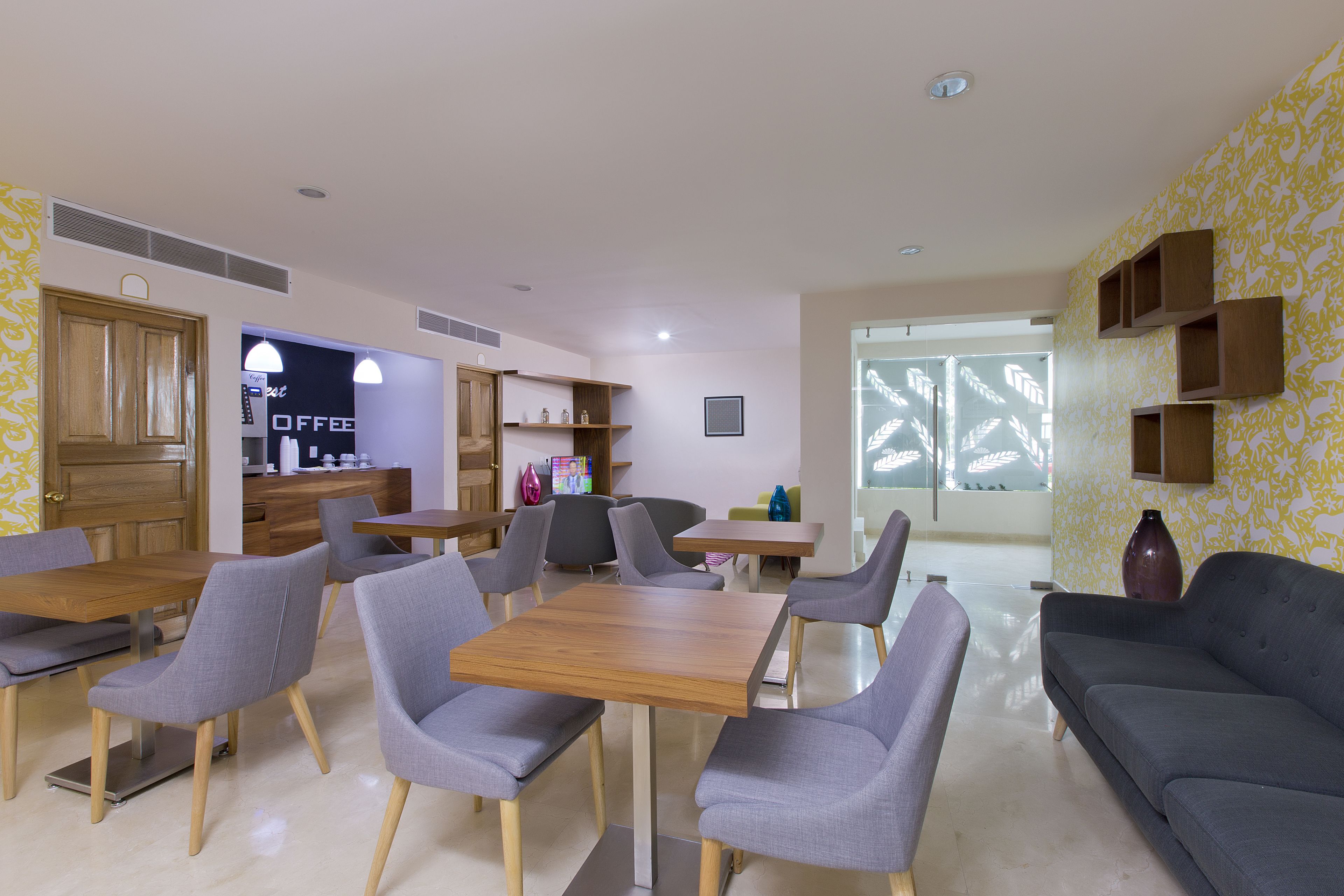 Un comedor moderno con mesas de madera y sillas tapizadas en gris, un sofá gris oscuro y una estación de café al fondo.