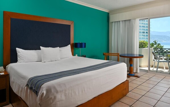 A hotel room with a large bed, a small table, and a balcony with a view.