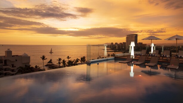 Ein Pool ohne Ende mit Liegestühlen und Sonnenschirmen, mit Blick auf einen Sonnenuntergang über dem Meer und einer Stadtkulisse im Hintergrund.