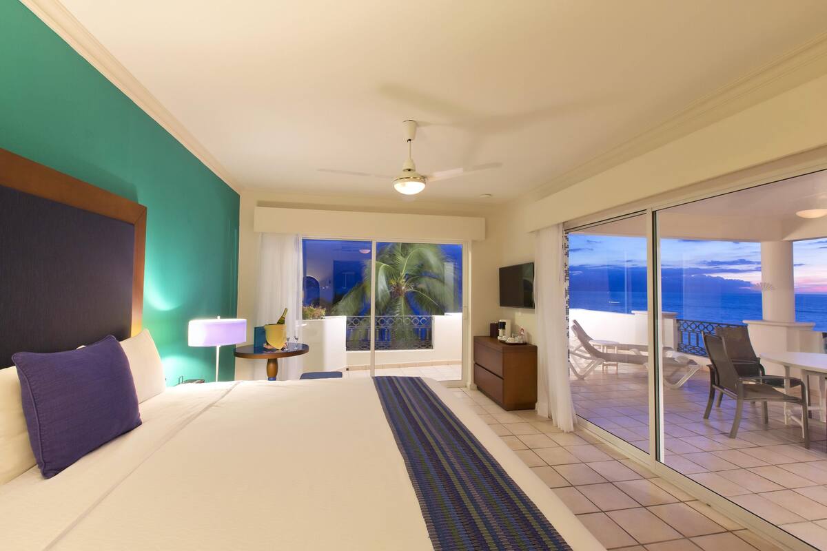 Une chambre d’hôtel avec un lit, une table de chevet, une lampe, une vue sur l’océan et un balcon équipé de chaises.