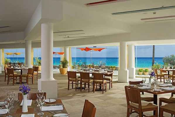 Un restaurante con mesas dispuestas para comer, grandes ventanales con vistas al océano y plantas en macetas.