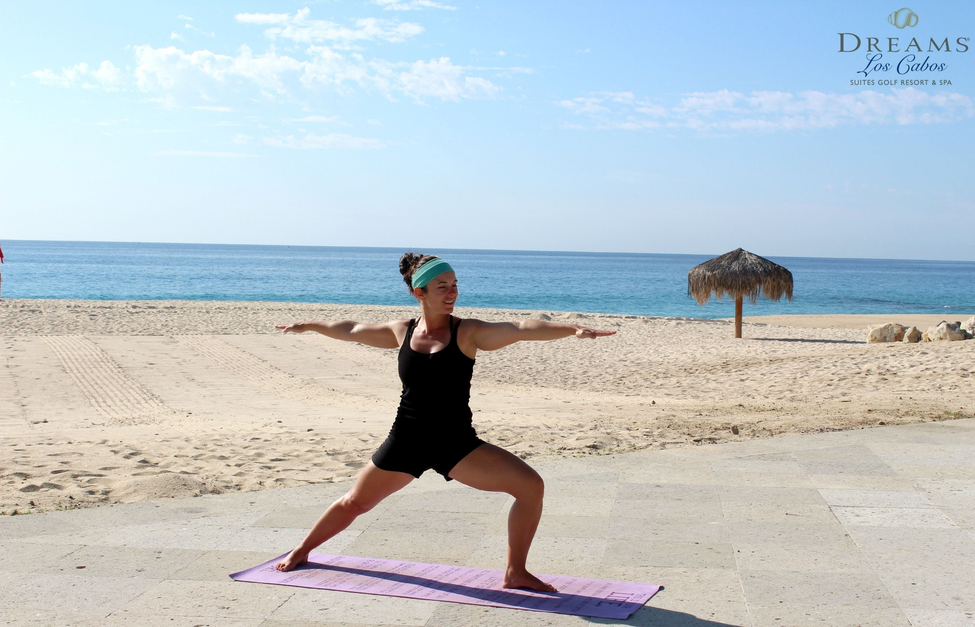 Une personne effectue une posture de yoga sur un tapis, sur une plage, sous un parasol en chaume, avec une vue dégagée sur l’océan.