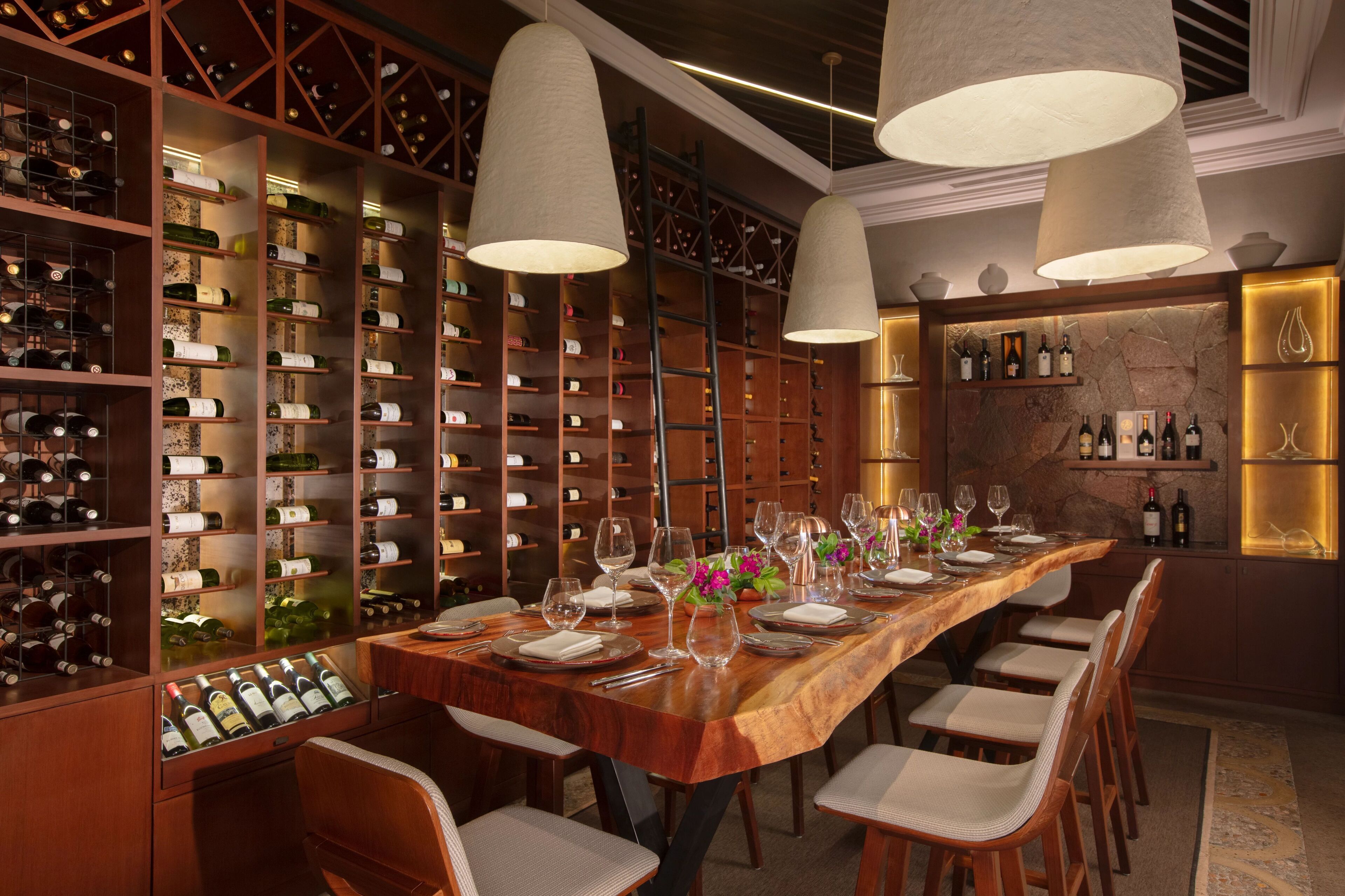 A wine cellar with wooden shelves and hanging wine racks, a long dining table set for six, and a bar area with a stone wall and shelves of bottles.