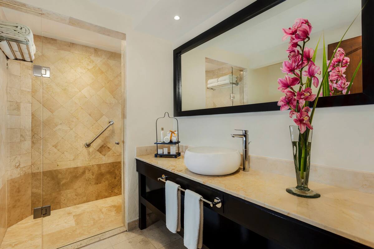 Een moderne badkamer met een douche met glazen wand, een grote spiegel en een vaas met roze bloemen op de wastafel.