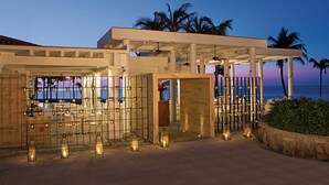 Un hotel frente a la playa con un espacio exterior cubierto, palmeras y vista al océano.