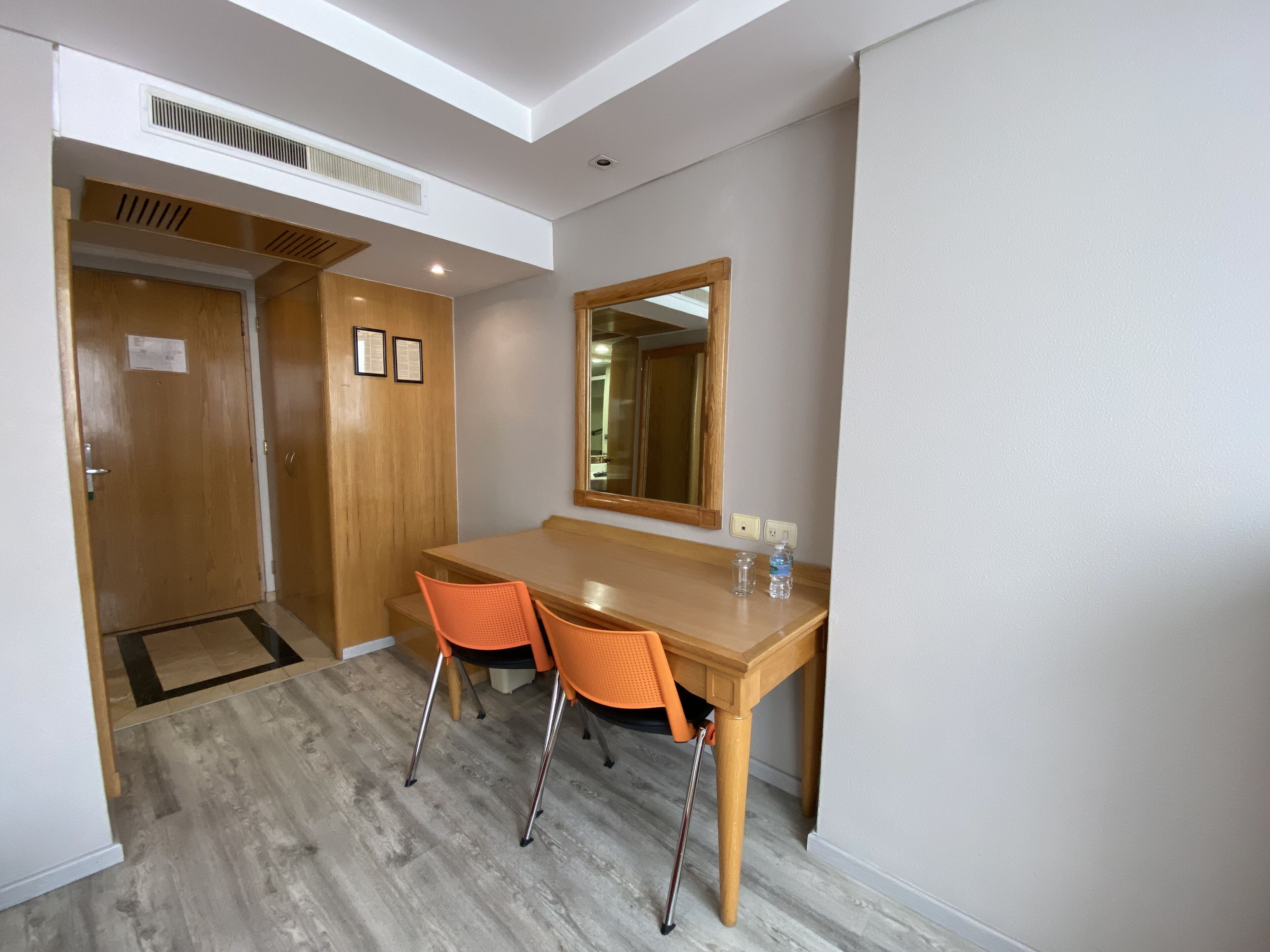 A hotel room with a wooden desk, two orange chairs, a large mirror, and a wooden door.