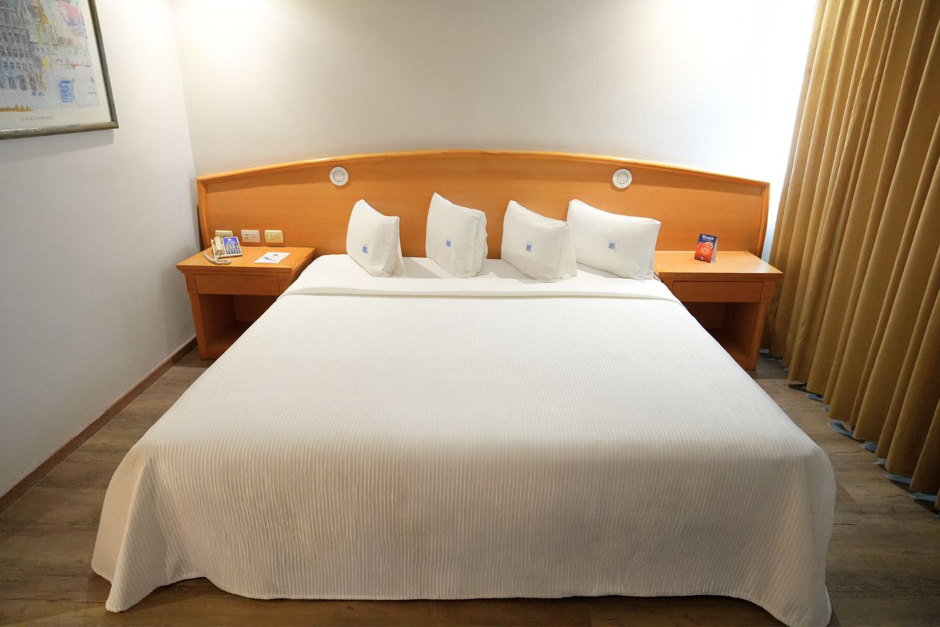 Ein Hotelzimmer mit einem großen Bett, einem hölzernen Kopfteil, zwei Nachttischen mit Telefon und Buch sowie einem Vorhang auf der rechten Seite.