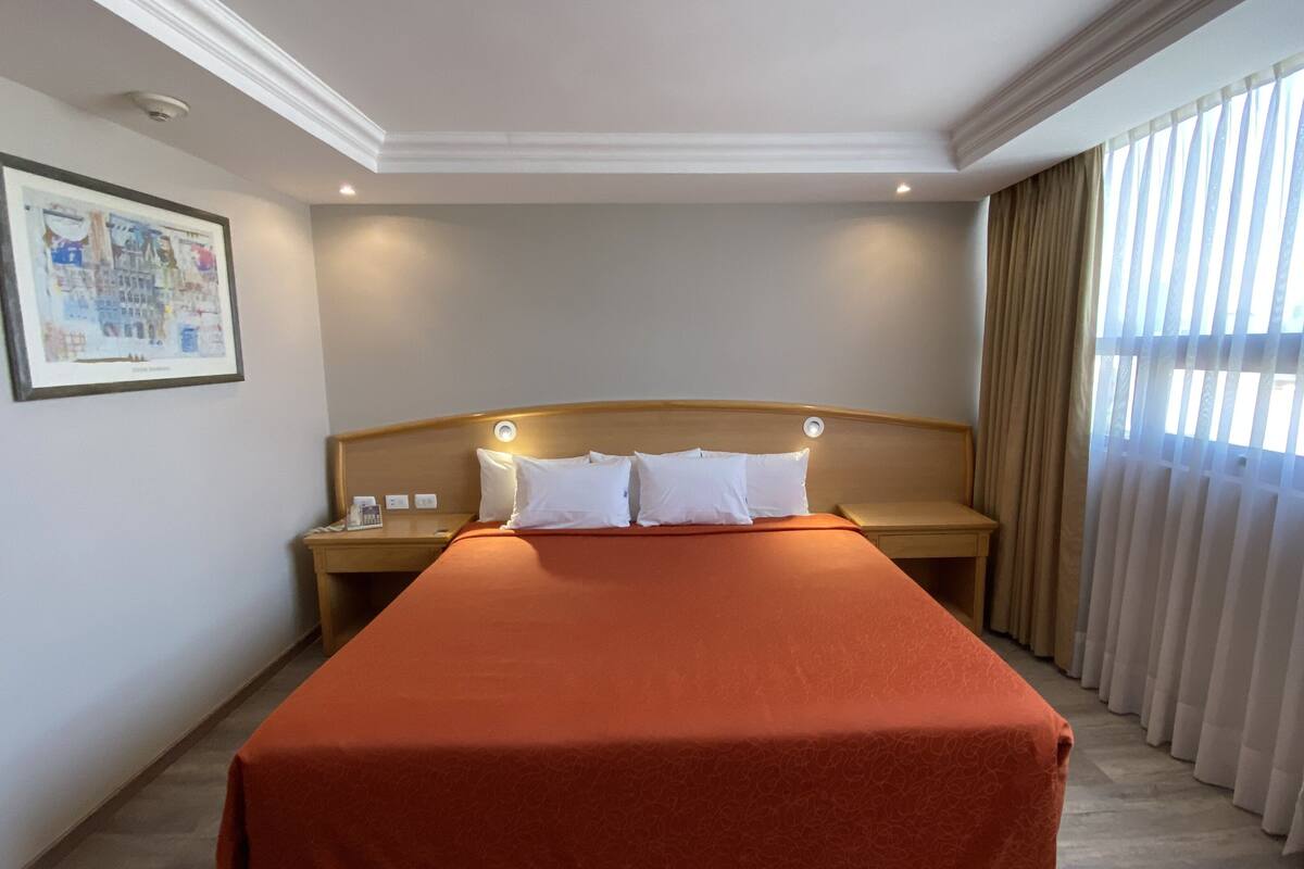 A hotel room with a large bed, a nightstand, a wall-mounted clock, and a window with curtains.