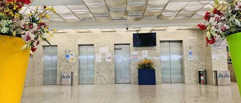 A lobby with a tiled floor, two elevators, a reception desk, and decorative floral arrangements.