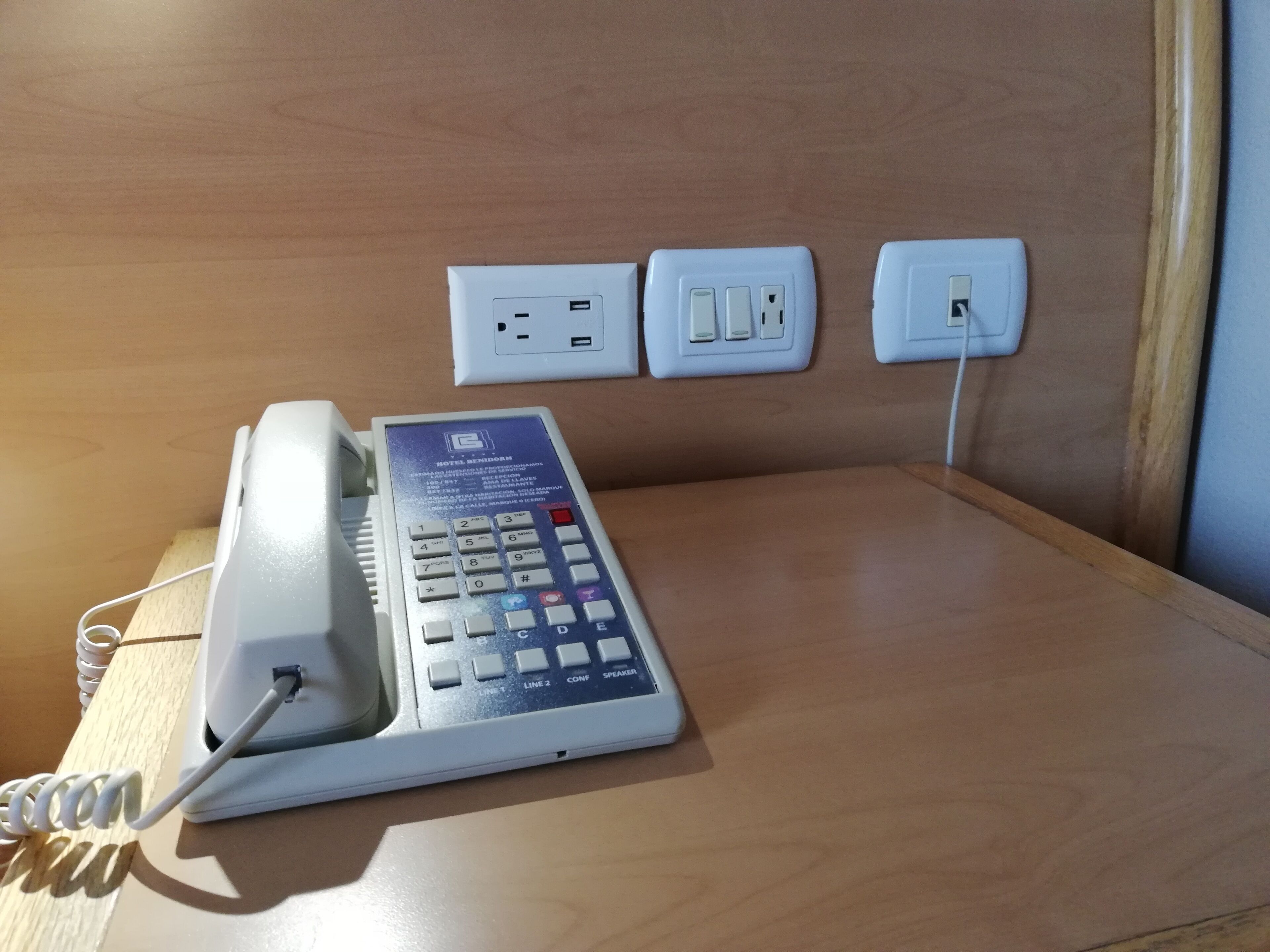 A hotel room with a desk, a telephone, and three electrical outlets on the wall.