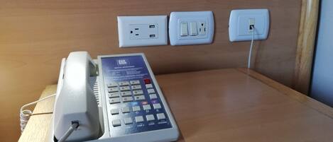 A hotel room with a desk, a telephone, and three electrical outlets on the wall.
