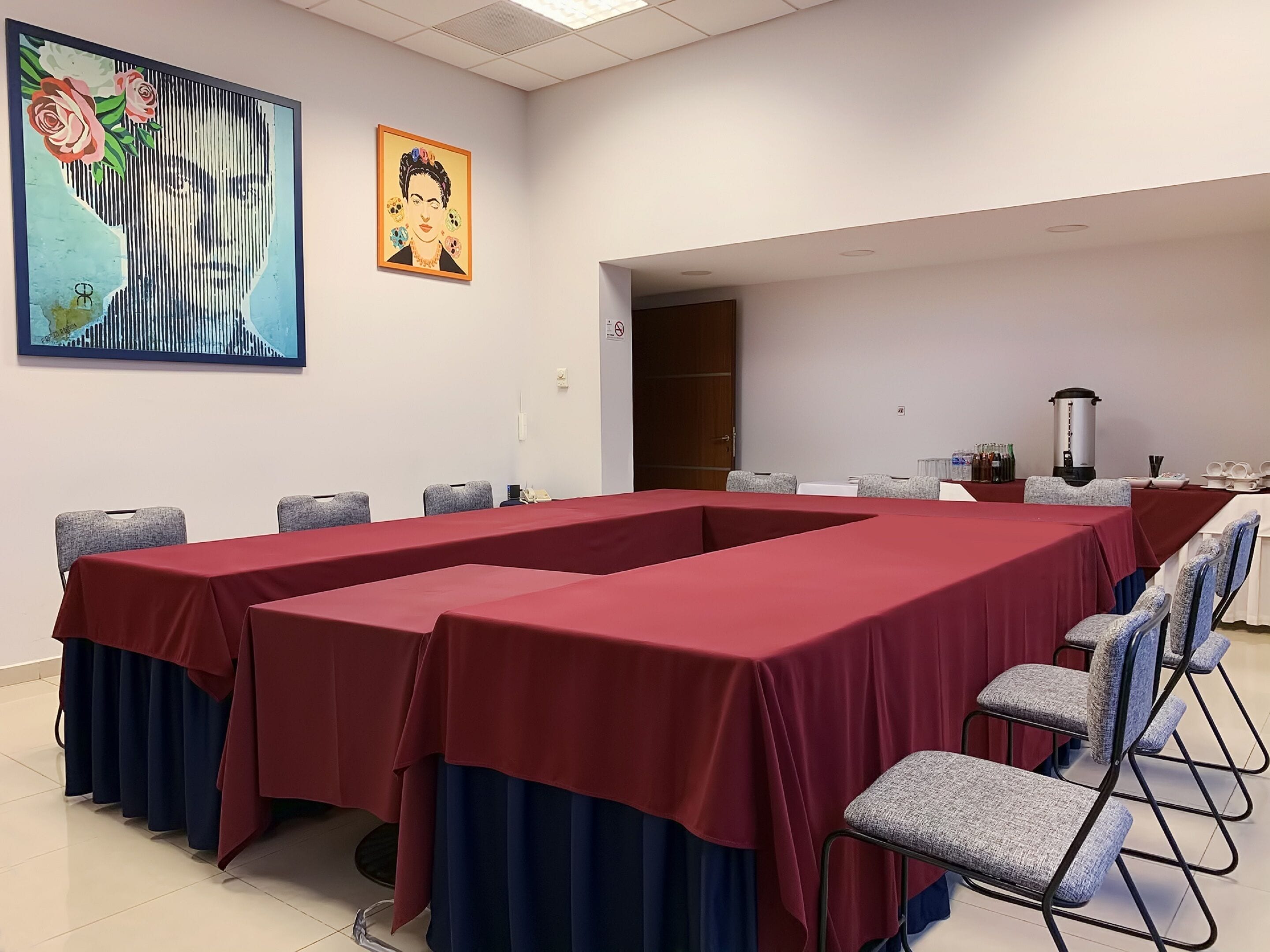 A conference room with a long table covered in red, surrounded by grey chairs. There are two paintings on the wall, one depicting a woman with flowers and the other a portrait.
