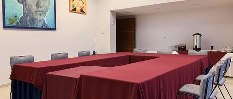 A conference room with a long table covered in red, surrounded by grey chairs. There are two paintings on the wall, one depicting a woman with flowers and the other a portrait.