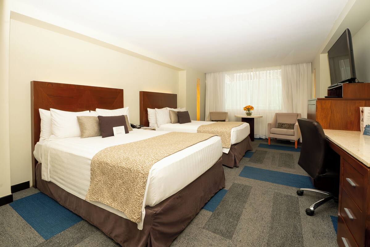 A hotel room with two beds, a desk, a chair, a television, and a small table with a vase of flowers.