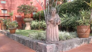 Un jardín con una estatua, plantas en macetas y un edificio rojo al fondo.