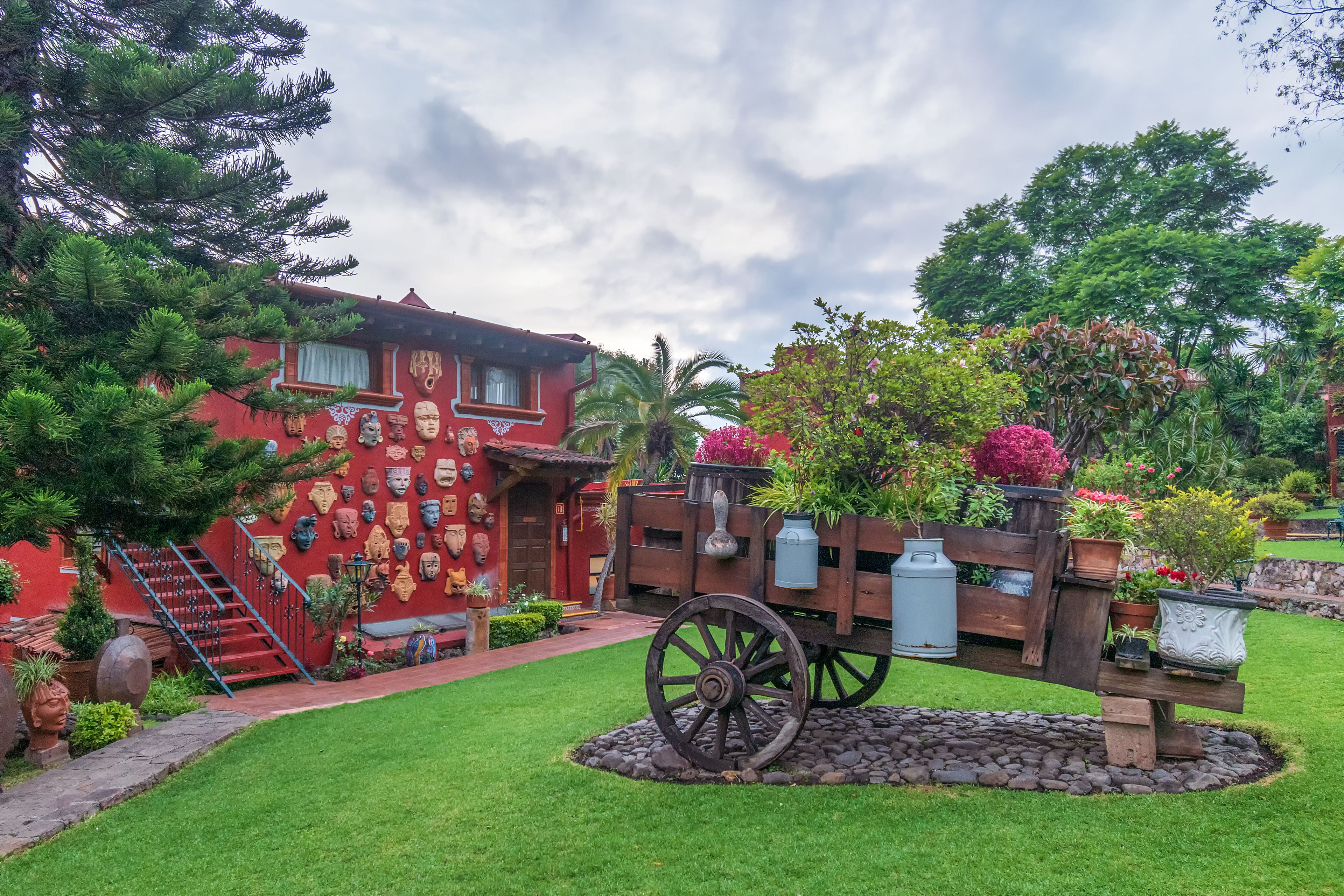 A red building with a mural of masks, a wooden fence with potted plants, and a stone pathway leading to a staircase.