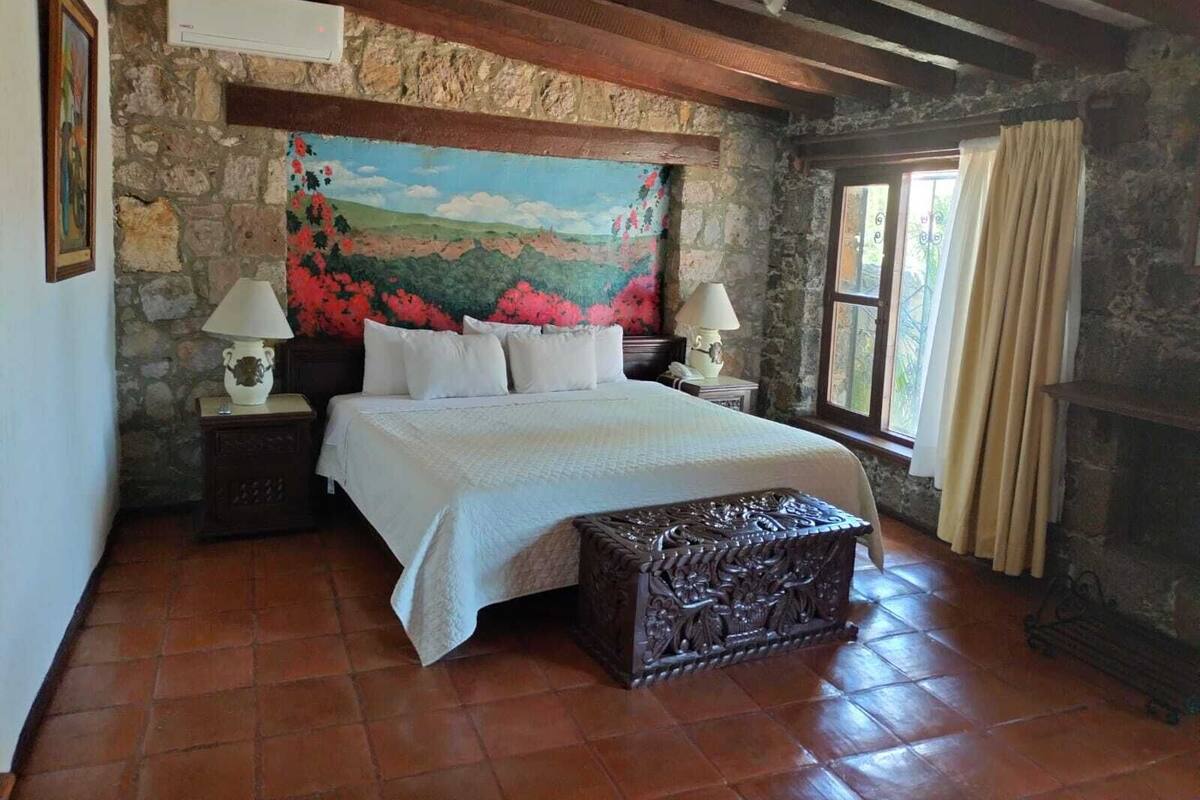 Une chambre avec un mur en pierre, un lit avec une literie blanche, deux tables de chevet avec des lampes, un grand tableau et une fenêtre avec des rideaux.