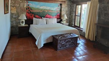 A bedroom with a stone wall, a bed with white bedding, two bedside tables with lamps, a large painting, and a window with curtains.