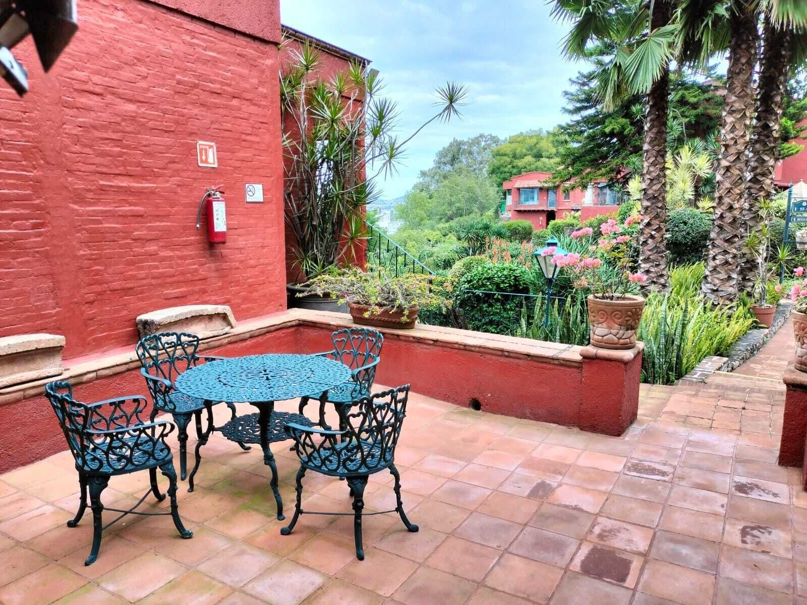 Un patio con una mesa redonda y cuatro sillas, rodeado de paredes rojas y plantas en macetas.