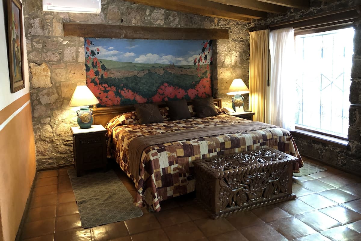 Une chambre avec un mur en pierre, un grand lit recouvert d’une courtepointe, deux tables de chevet avec des lampes, un banc en bois et une fenêtre avec des rideaux.