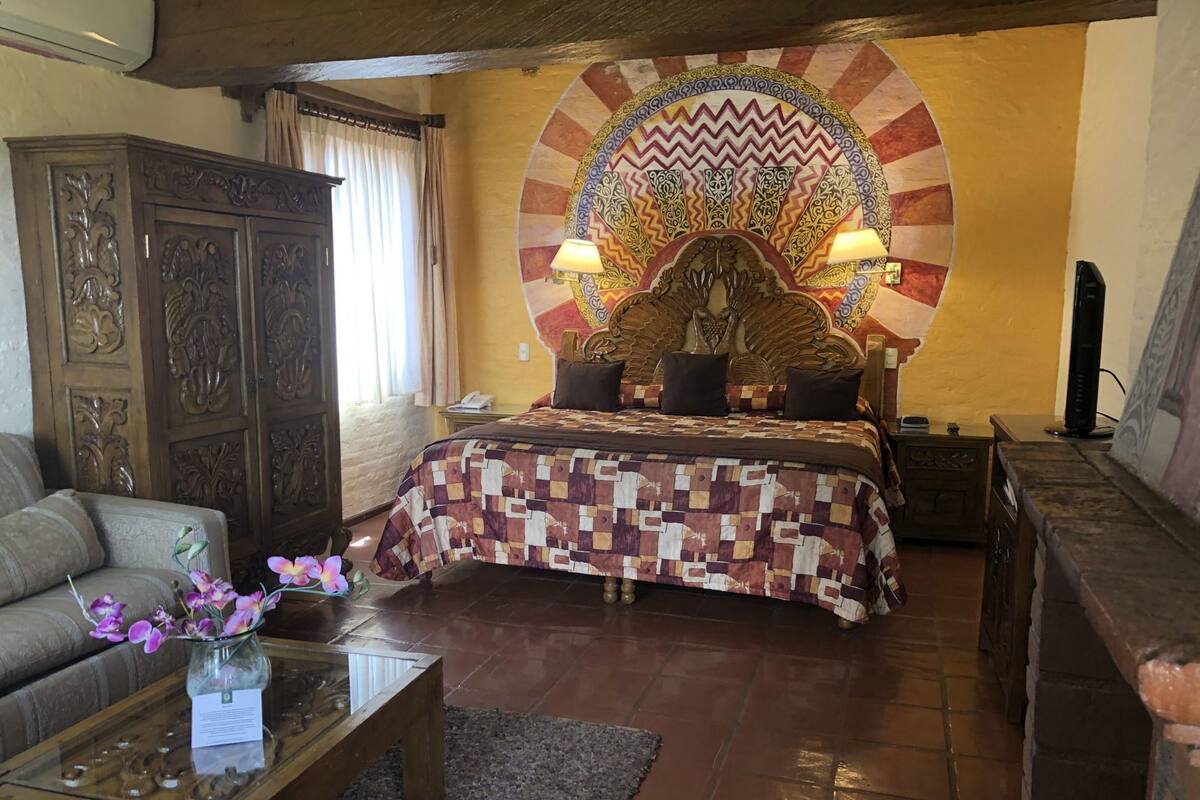 Une chambre à coucher avec une tête de lit à motifs, un lit recouvert d’une courtepointe, une armoire en bois, un canapé, une table basse ornée de fleurs et un téléviseur.