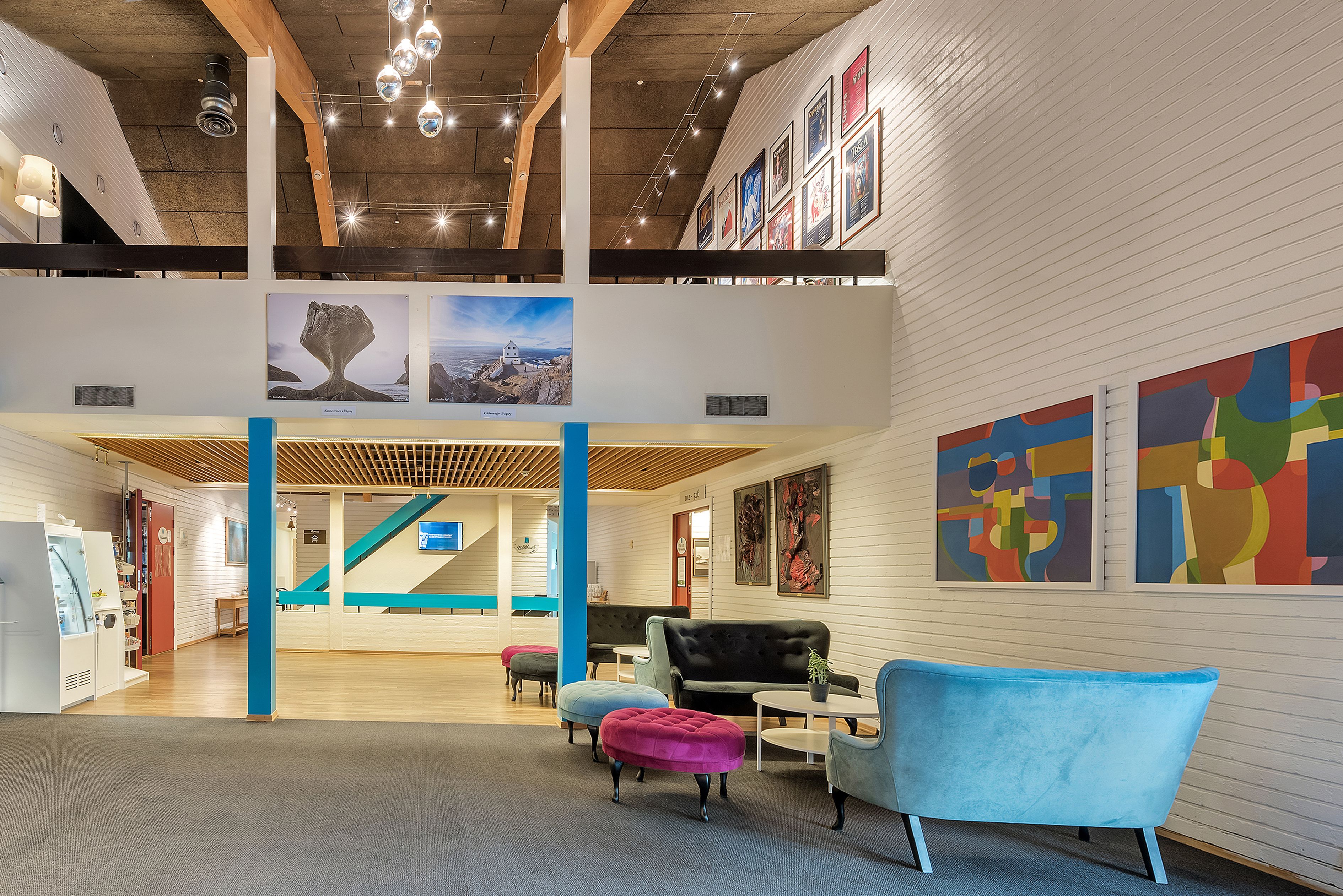A spacious lobby with a high ceiling, wooden beams, and a staircase. There are colorful artworks on the walls and a seating area with ottomans and a sofa.