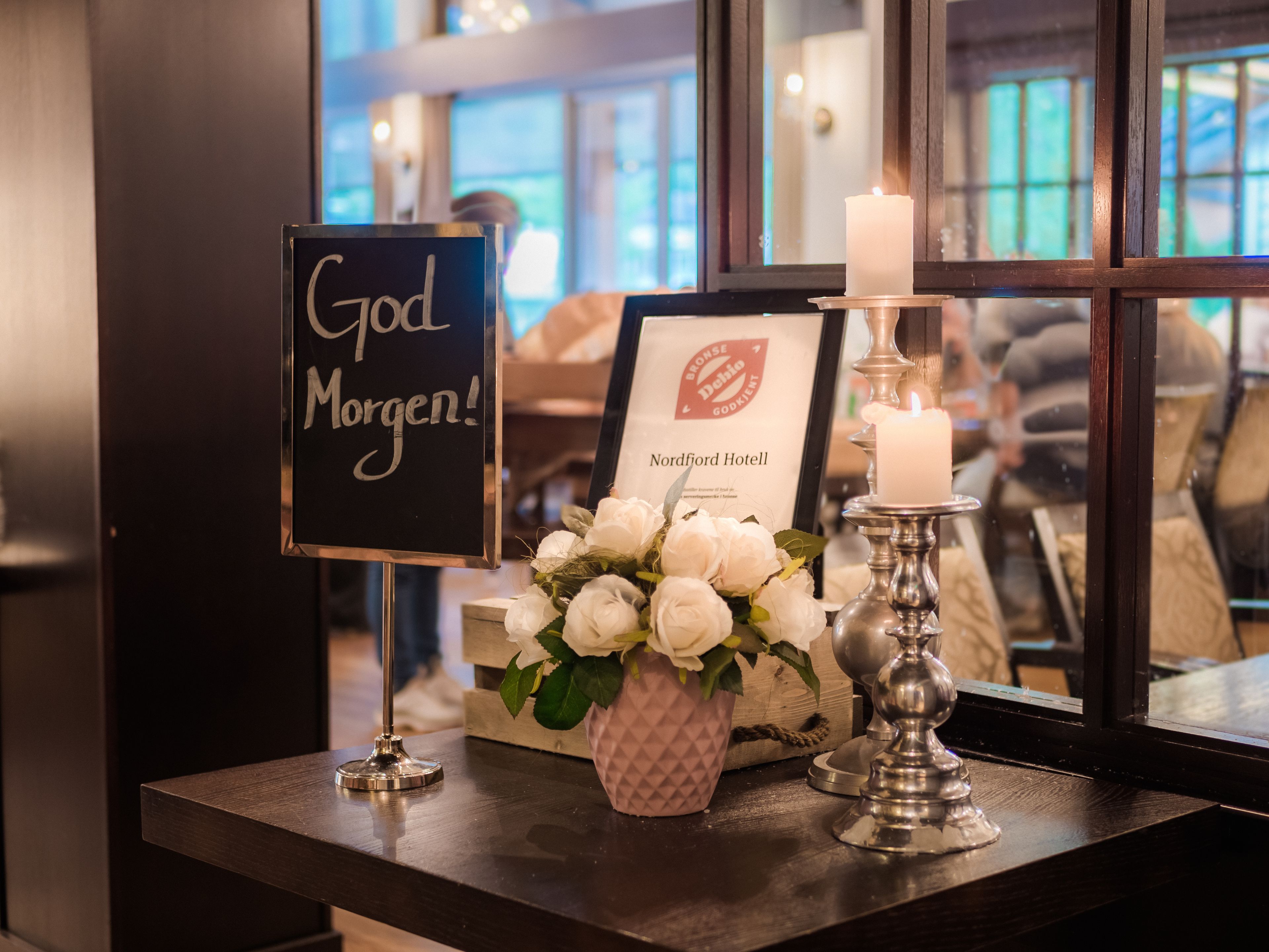 A table with a sign that reads "God Morgen!", a framed certificate, a candle, and a vase of white flowers.