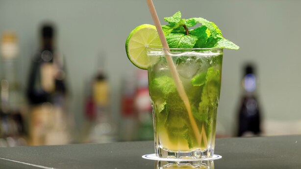 Un vaso de una bebida verde con una rodaja de lima y hojas de menta, sobre una superficie reflectante.