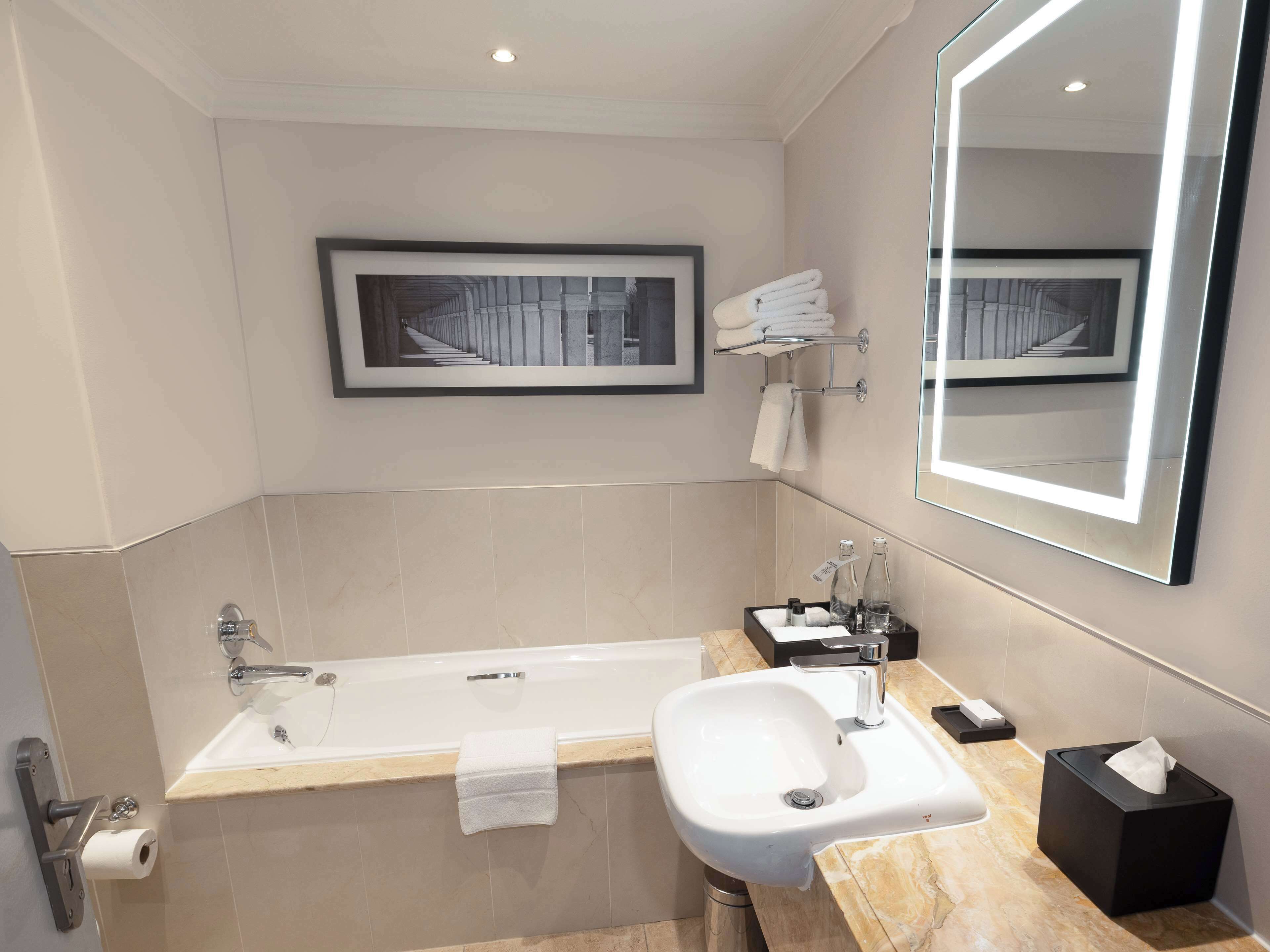 Un baño moderno con bañera, lavabo y un espejo grande. Hay un cuadro en la pared y toallas en un perchero.