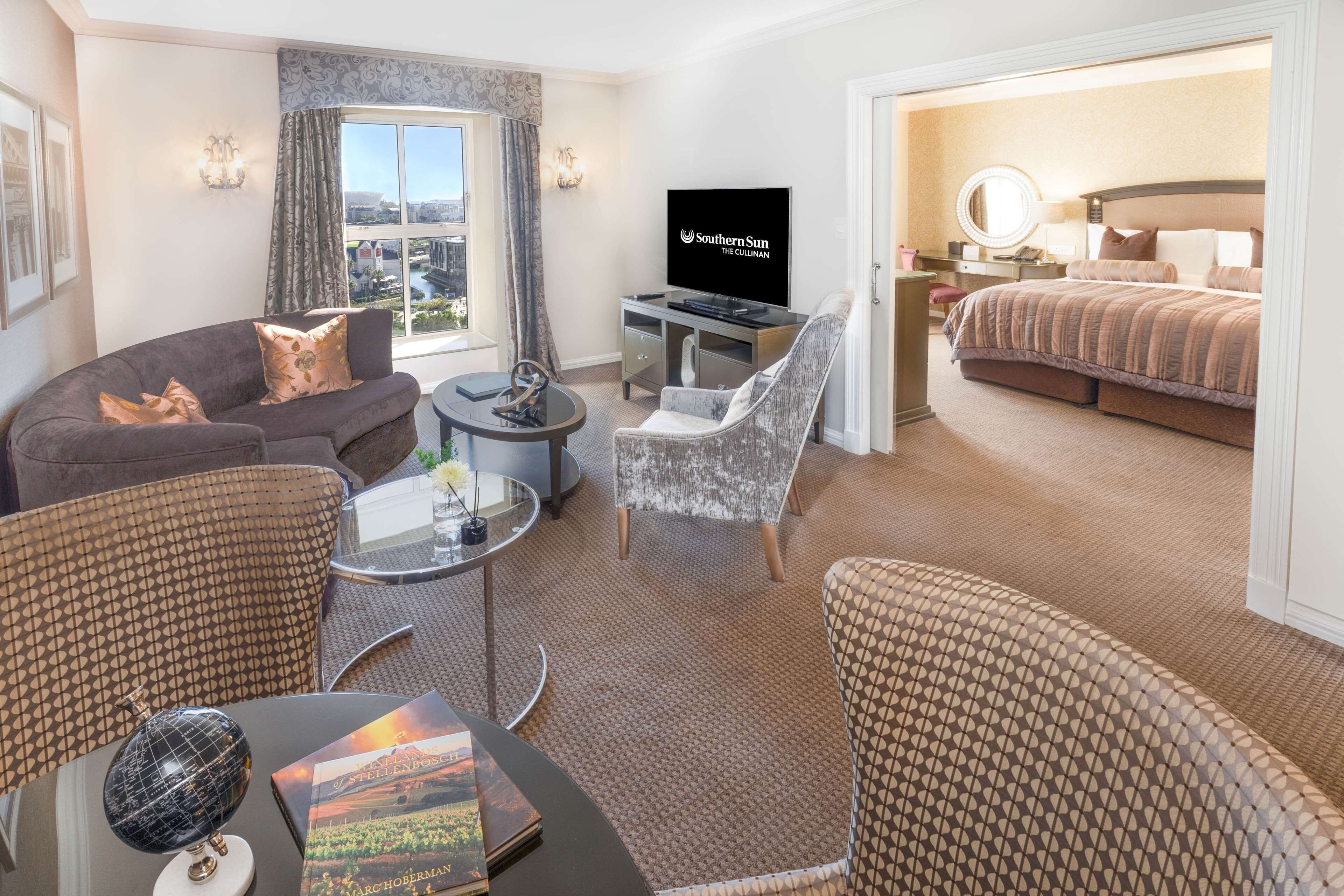 A hotel room with a sofa, armchair, and a bed. There's a TV on a stand, a small table with a globe and a book, and a window with curtains.
