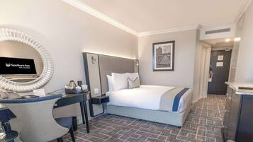 A hotel room with a bed, a desk with a computer, a chair, a mirror, and a framed picture on the wall.