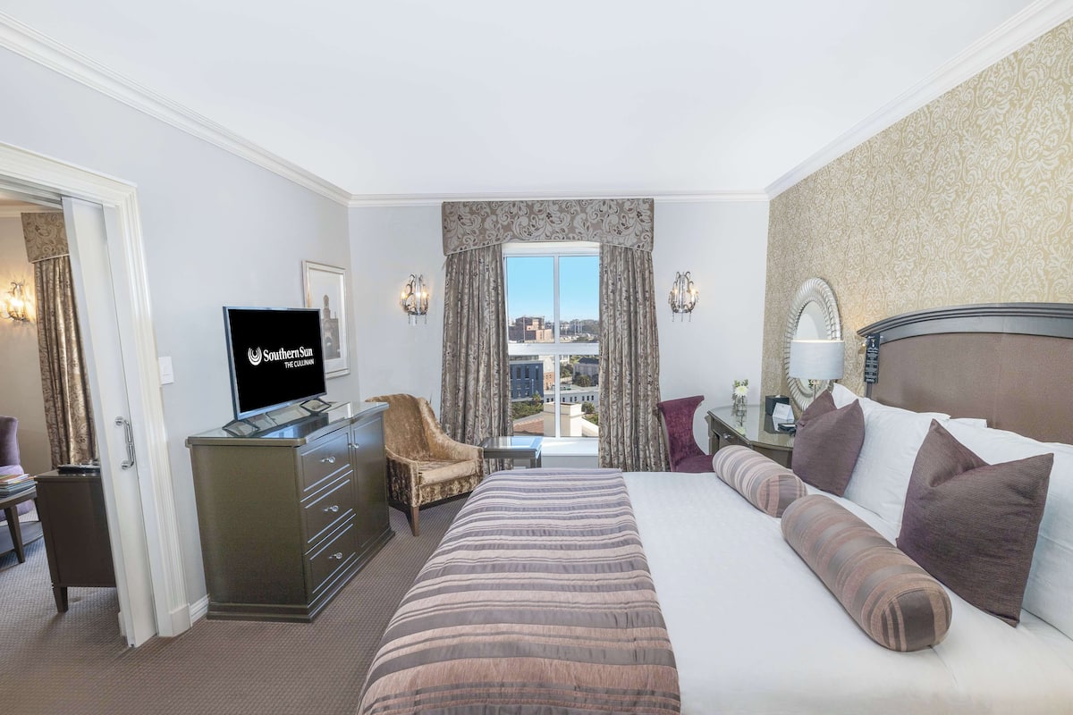 A hotel room with a large bed, a desk with a TV, a chair, and a view of the city through the window.