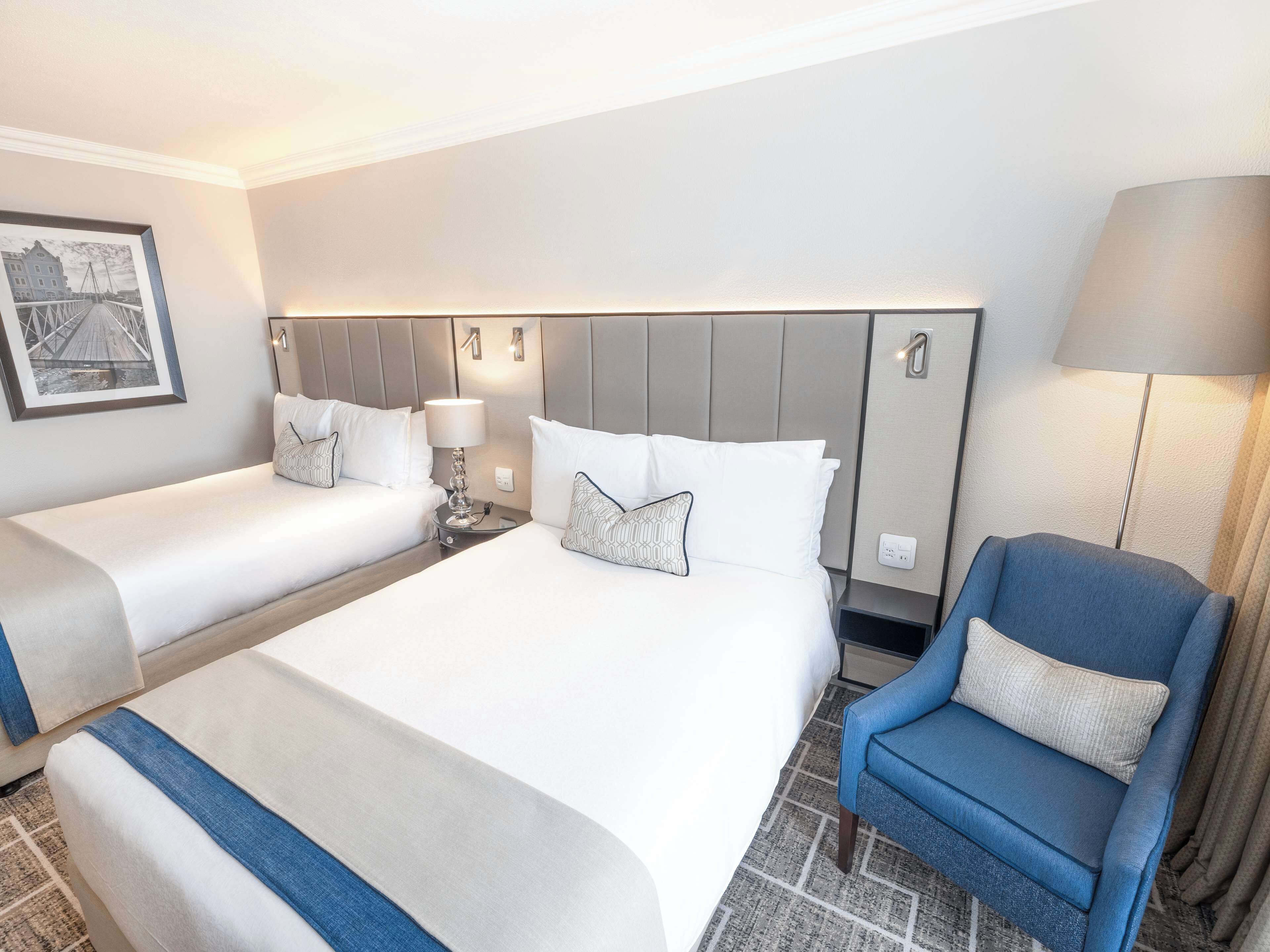 Quarto de hotel com duas camas, uma poltrona azul e uma luminária fixada na parede.
