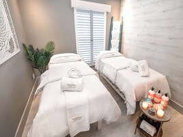 A spa room with two beds, each with a white towel and pillow labeled "Mangwansari Bath Spa." There are candles and bottles on a small table beside the beds.