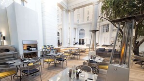 An outdoor dining area with tables and chairs, a pizza oven, and a large window overlooking a building. - Southern Sun The Cullinan (Cape Town)
